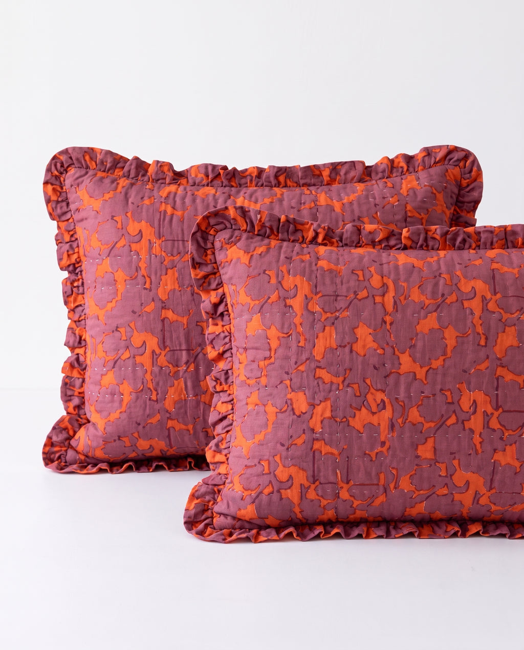 Two rectangular decorative pillows with ruffled edges, featuring an abstract orange pattern on a muted mauve-purple background, set against a plain white studio background.
