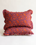Two ruffled decorative pillows stacked on a light neutral background. The pillows feature a textured, quilted pattern in a deep plum purple and bright orange.