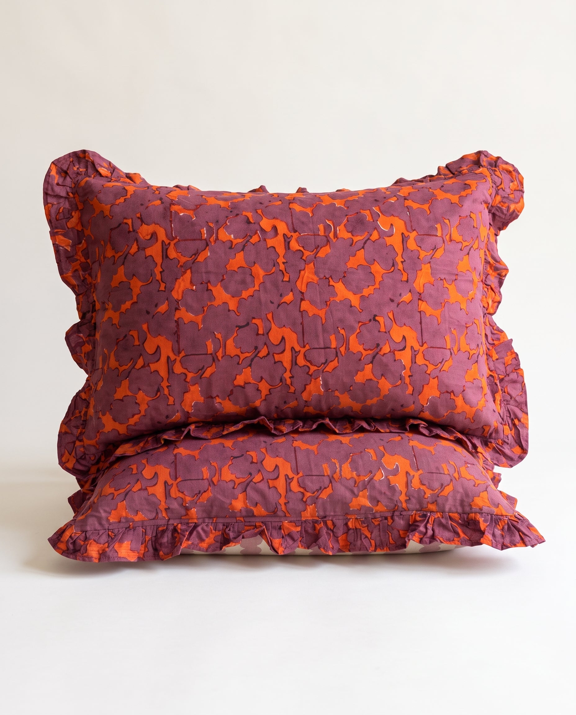Two ruffled decorative pillows stacked on a light neutral background. The pillows feature a textured, quilted pattern in a deep plum purple and bright orange.