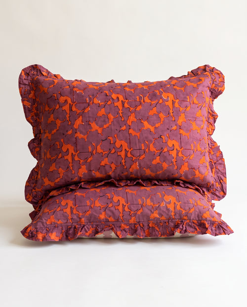 Two ruffled decorative pillows stacked on a light neutral background. The pillows feature a textured, quilted pattern in a deep plum purple and bright orange.