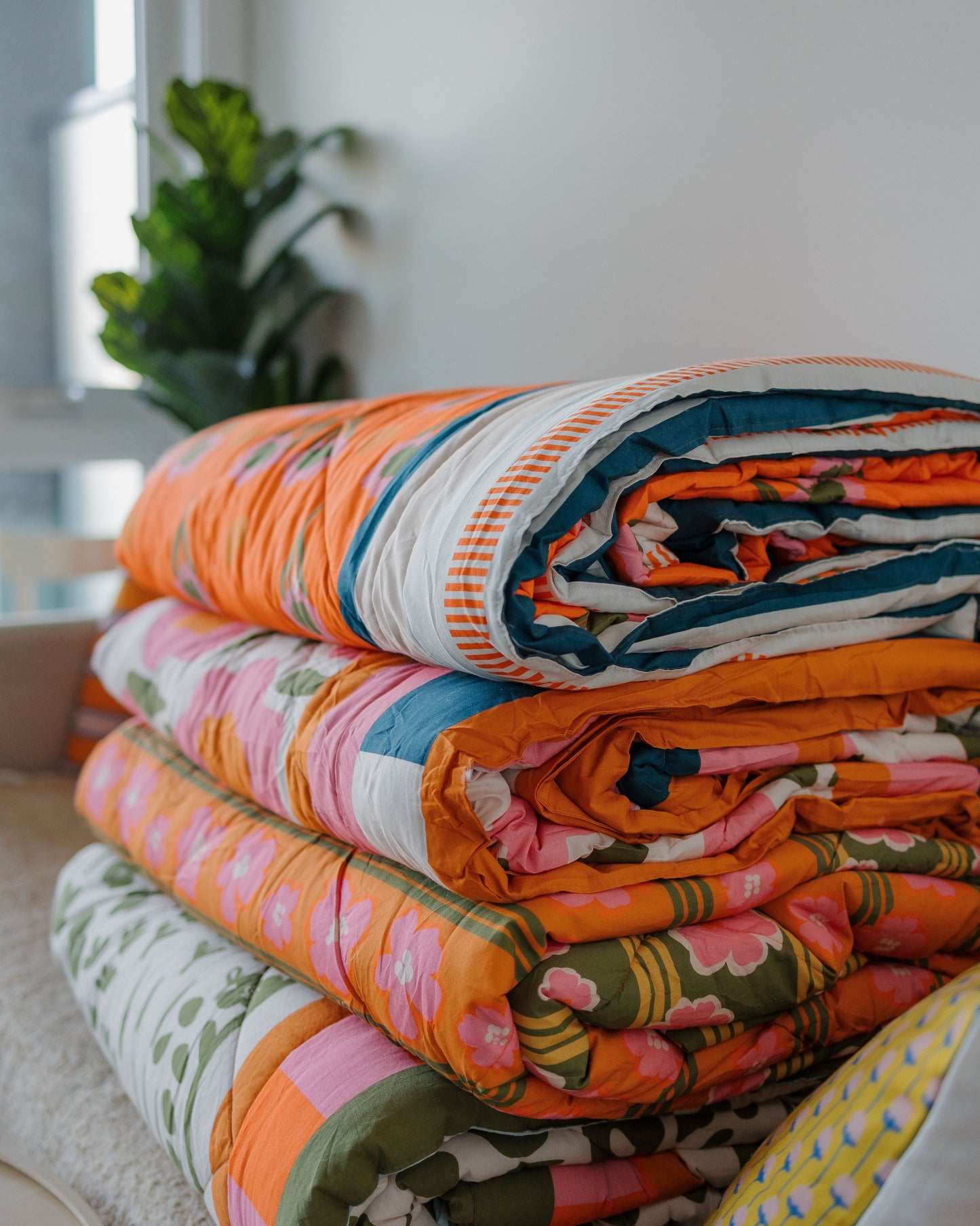 Stack of colorful patterned blankets on a bed with a blurred plant in the background