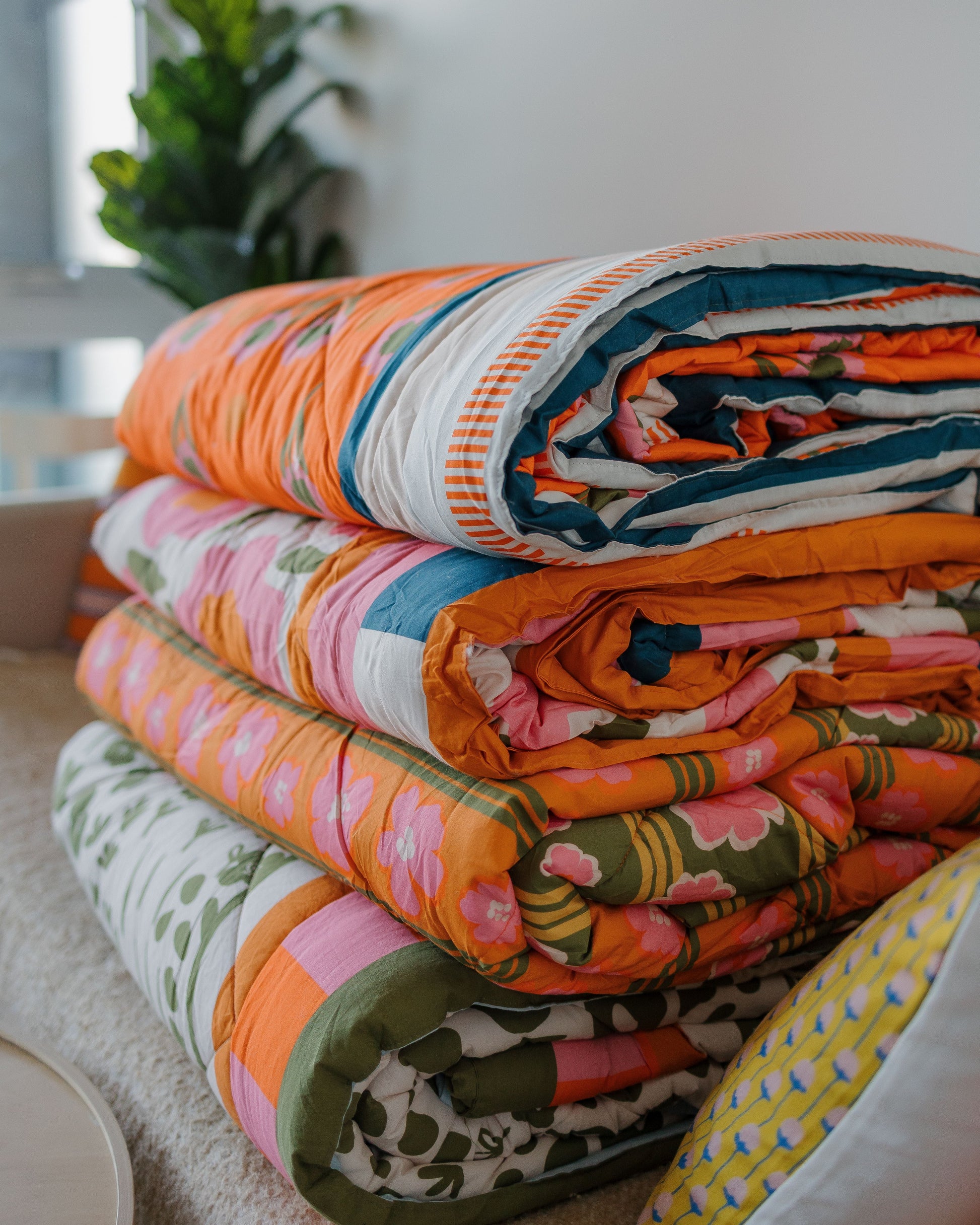 Stack of colorful patterned blankets on a bed with a blurred plant in the background