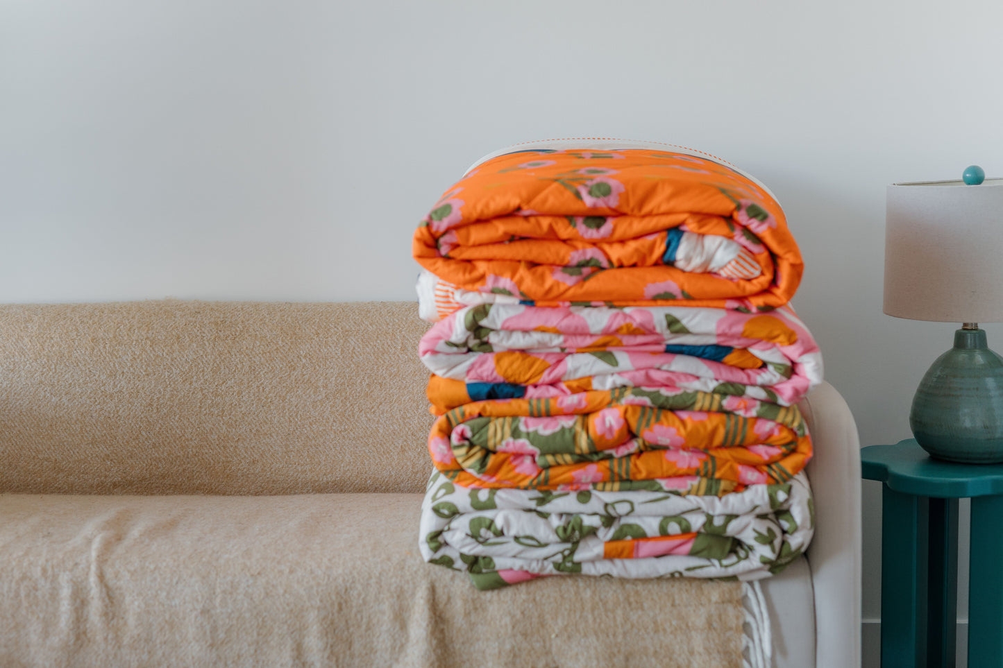 Stack of floral blankets on a beige couch with a lamp in the background