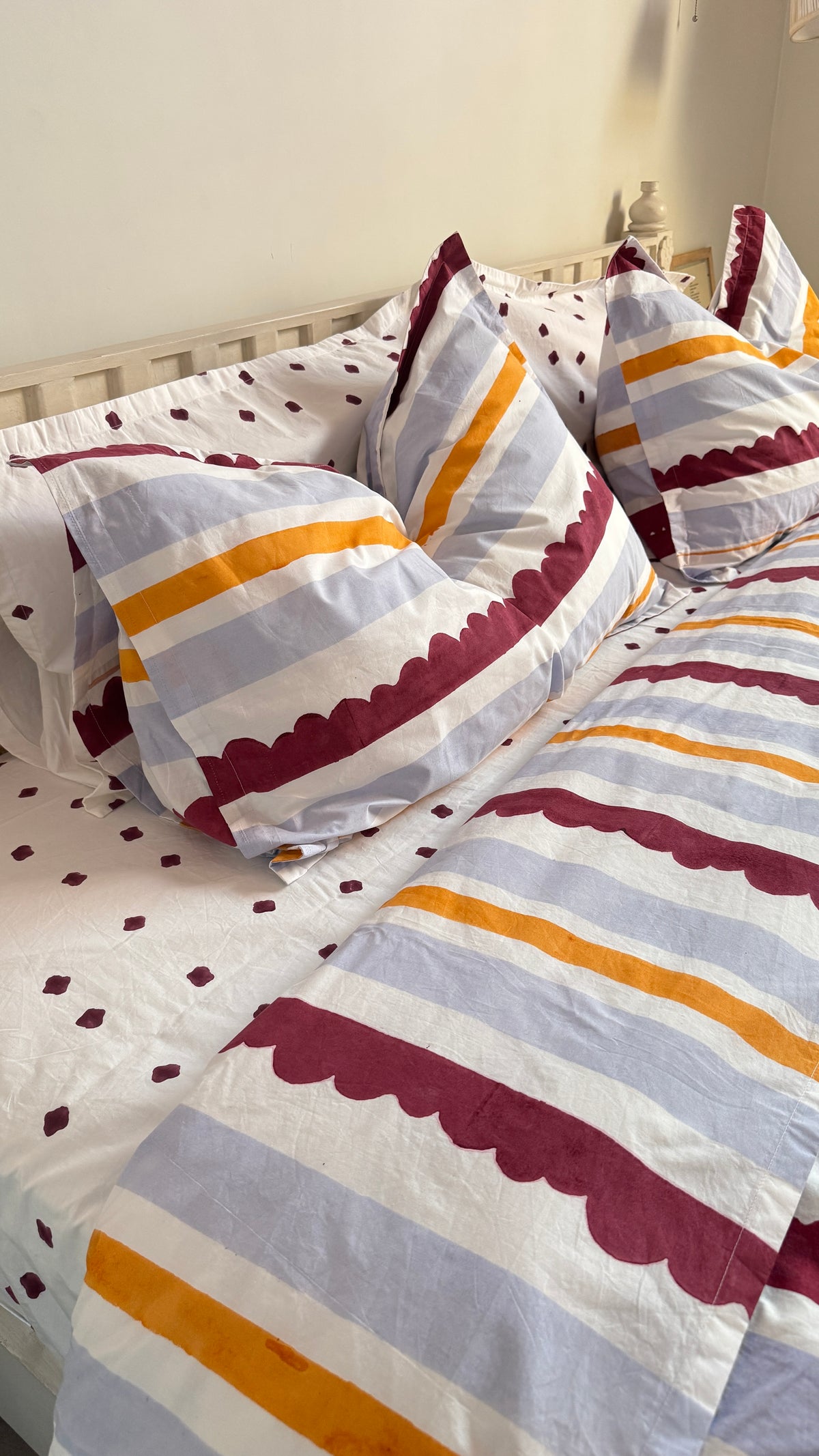 A close-up of a bed styled with a striped bedding set featuring white, dusty blue, mustard yellow, and burgundy stripes with scalloped edges, alongside matching polka-dot pillows.