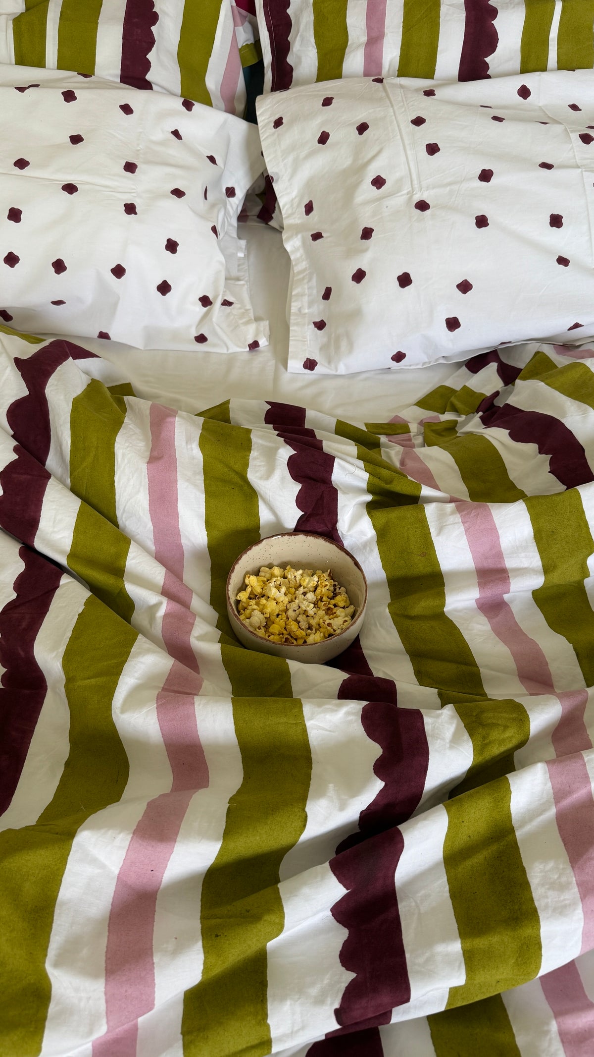 A cozy bed styled with a striped bedding set featuring olive green, dusty pink, and dark burgundy stripes, white pillows with burgundy diamond patterns, and a small bowl of popcorn in the center.