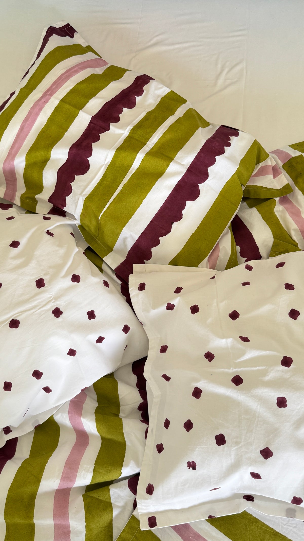 A close-up of a bedding set with white pillows and a striped duvet cover. The duvet features vertical stripes in olive green, white, and dusty pink with scalloped edges, while the pillows are white with small dark burgundy polka dots.