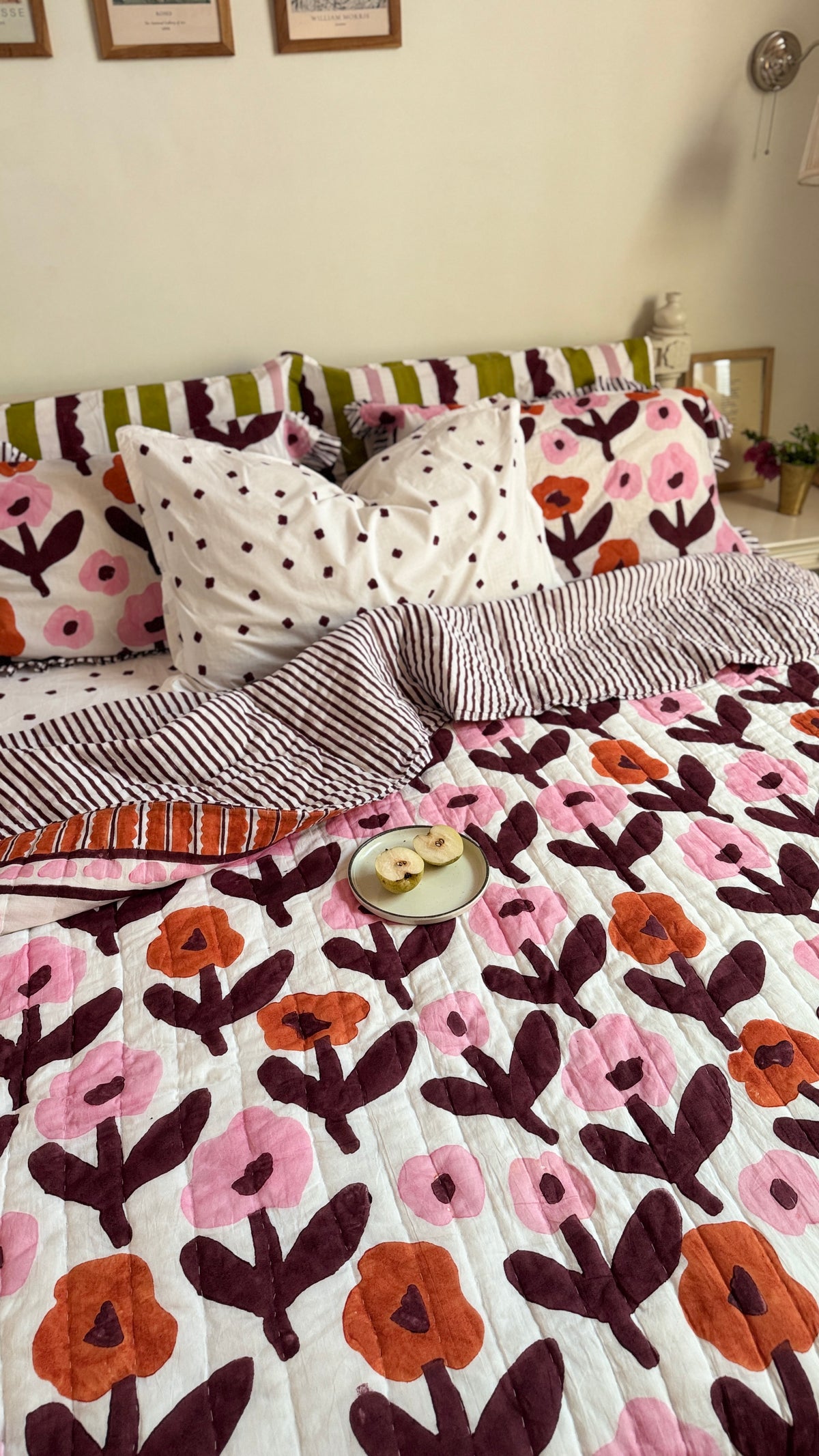 A cozy bed styled in a bedroom setting, featuring a quilted bedding set with a floral pattern in pink, orange, and dark brown on a white background. The bed is decorated with matching patterned pillows, including one with black polka dots on white, and a striped pillowcase.