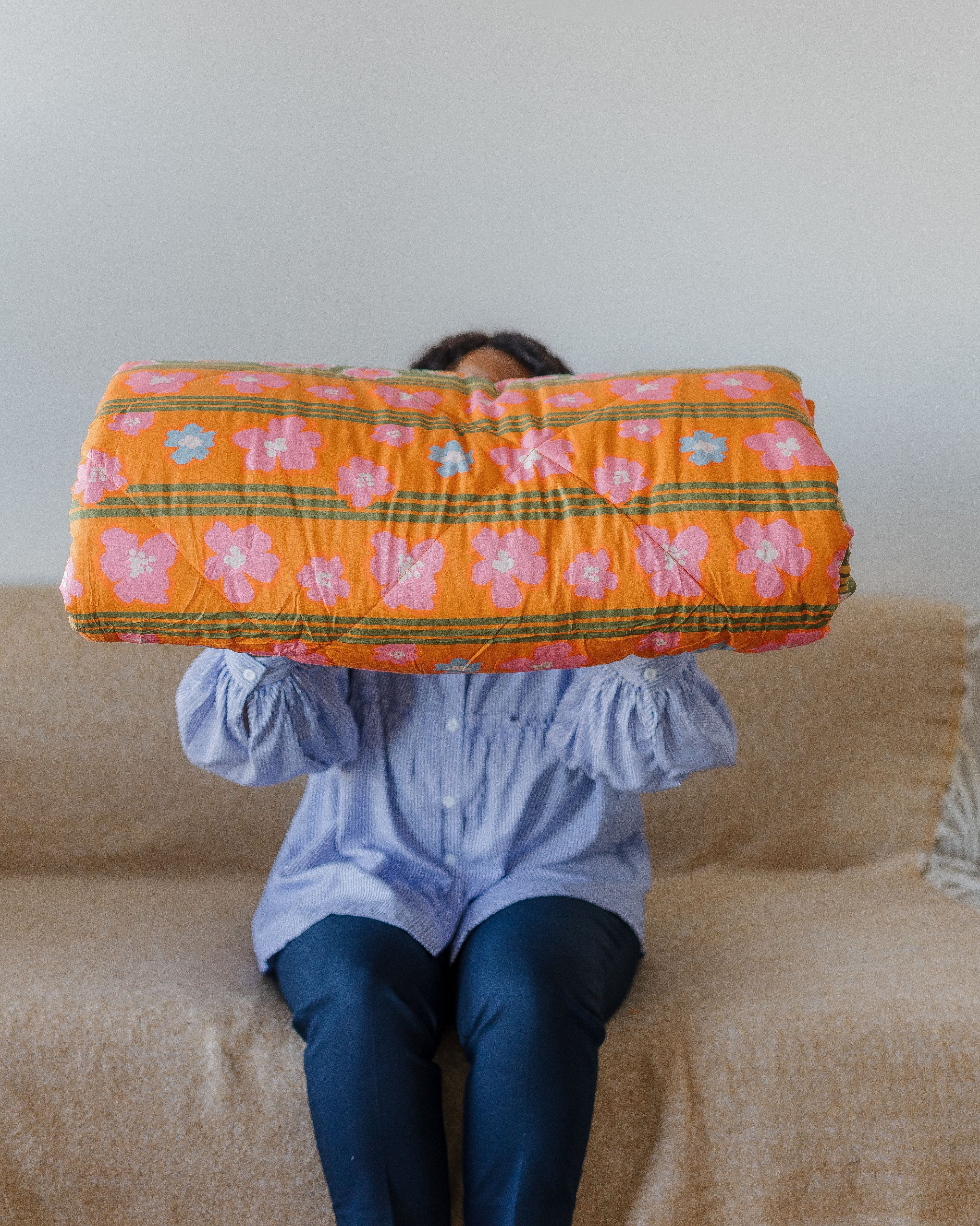 Person sitting on a couch holding an orange floral-patterned quilt blanket over their head.